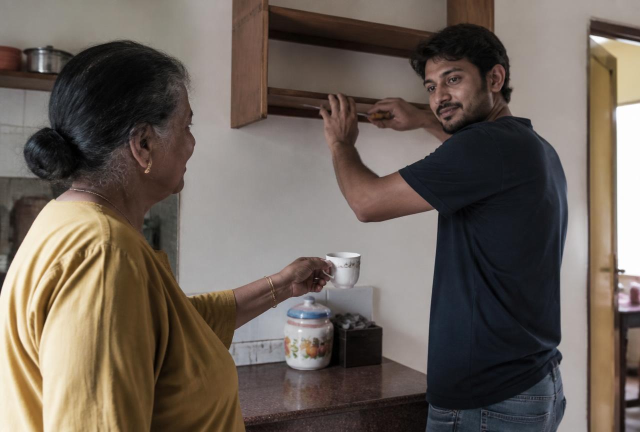 An older woman handing a cup of tea to a neighbour fixing a shelf in her kitchen